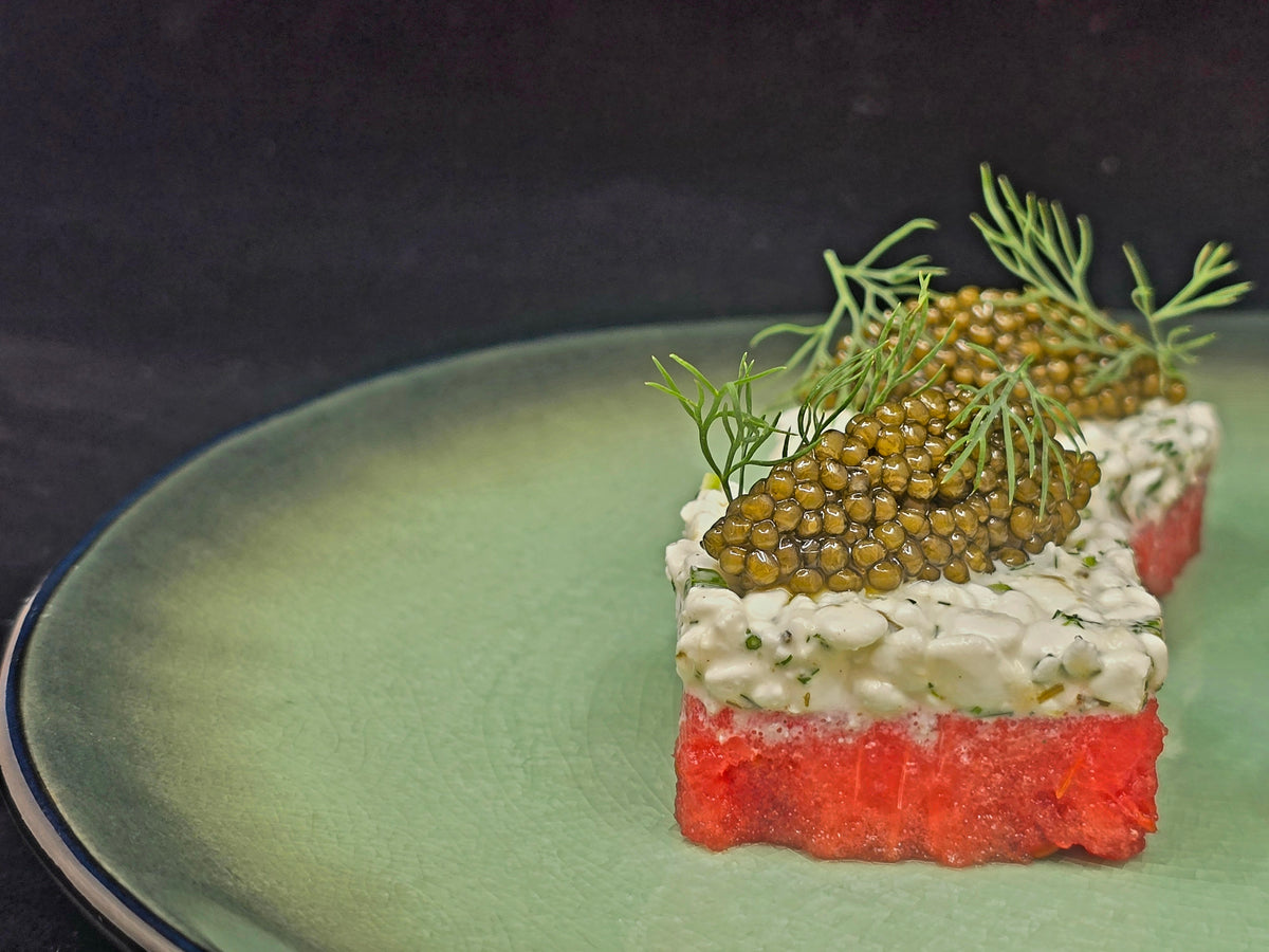 A close-up of a plated food item featuring a bright red watermelon base layer, a thick off-white creamy middle layer of horseradish, and a top of green caviar garnished with dill, served on a light green plate against a dark background.