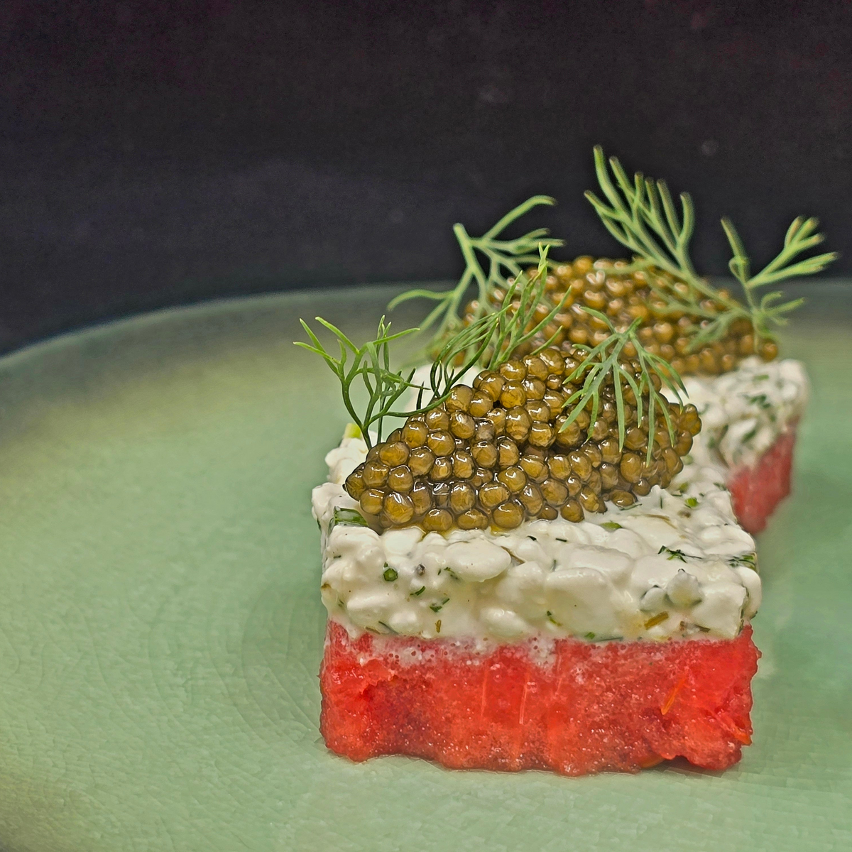 A close-up of a plated food item featuring a bright red base, a thick layer of white cream, and a top garnish of green caviar and dill on a light green plate.
