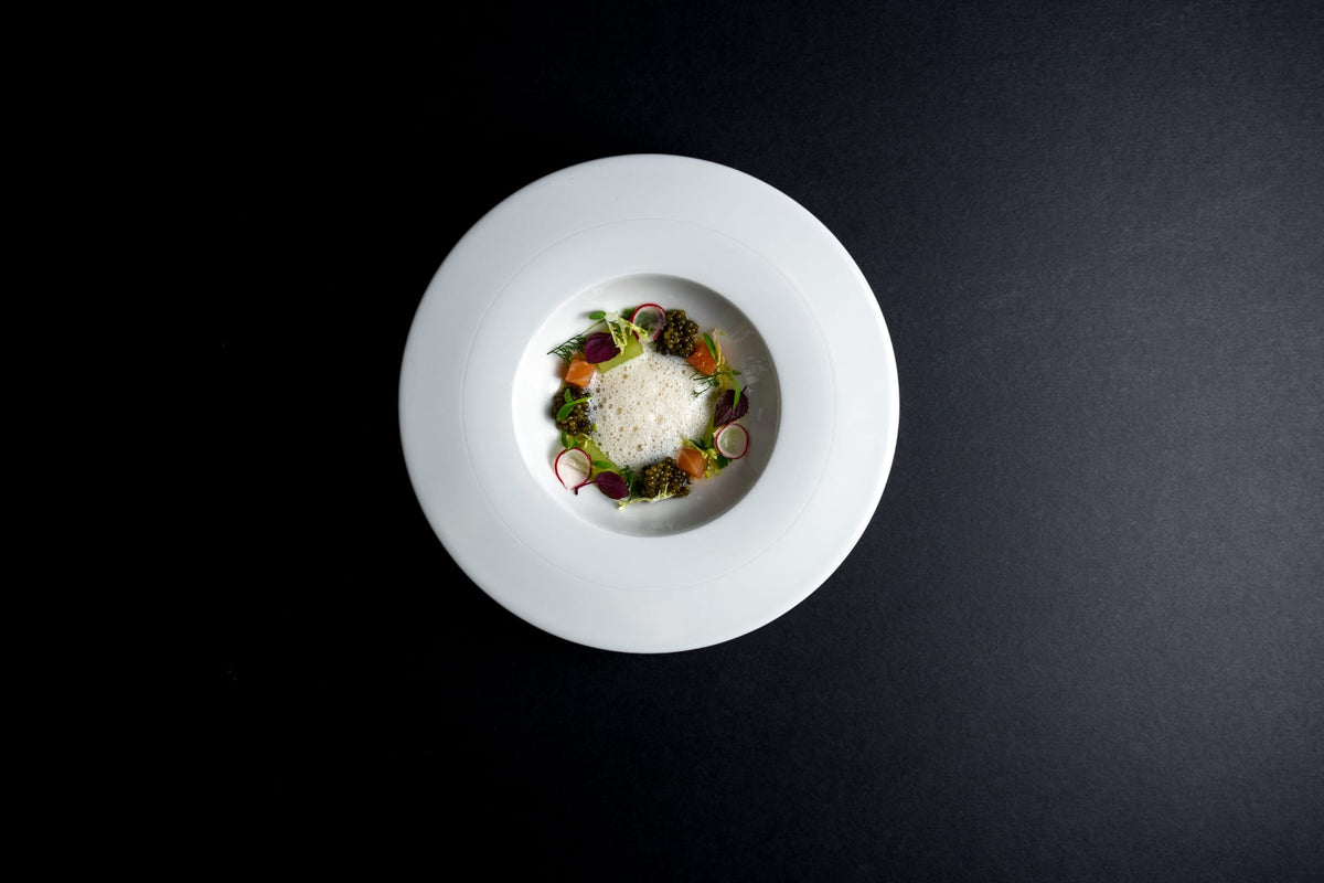 A top-down view of a white ceramic deep-dish plate containing a gourmet food dish, featuring a central mound of white cream or cheese surrounded by a ring of colorful vegetables and greens, set against a solid black background.