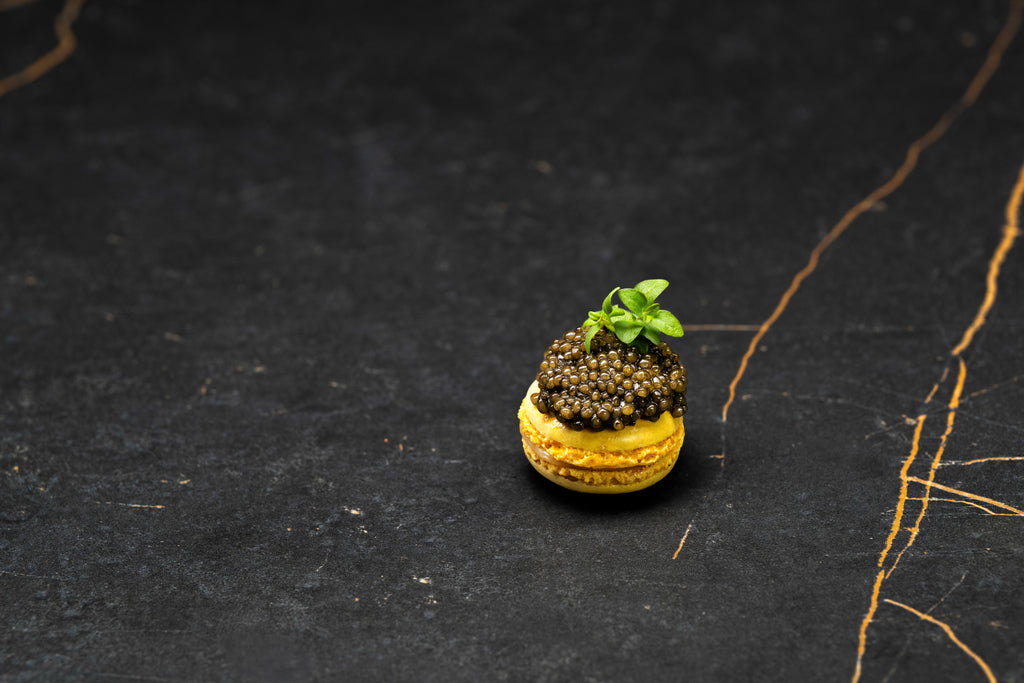 Yellow gourmet macaron topped with glossy dark caviar pearls and fresh green microgreens.