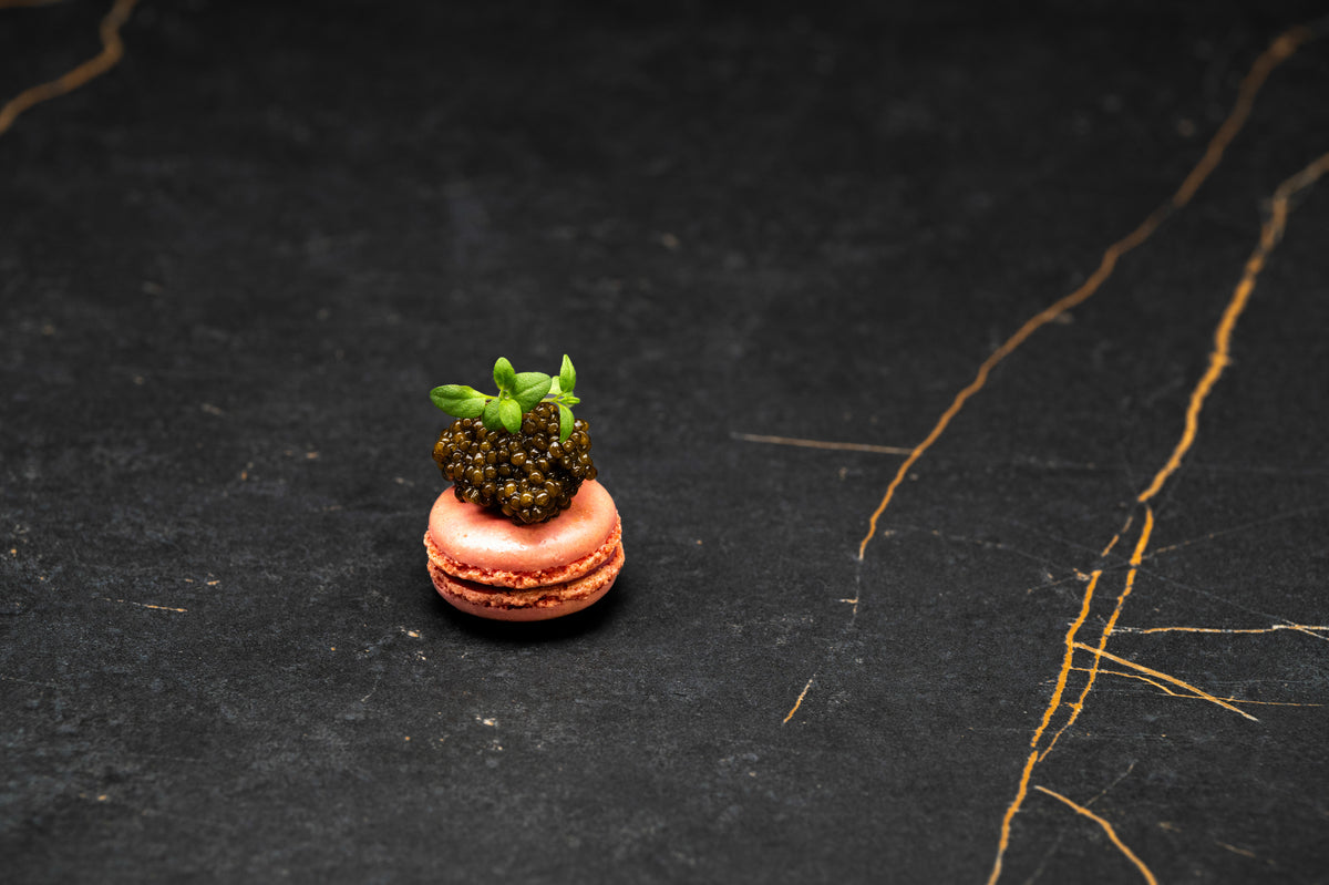 Pink macaron topped with a generous serving of dark caviar pearls and fresh microgreens on black marble.