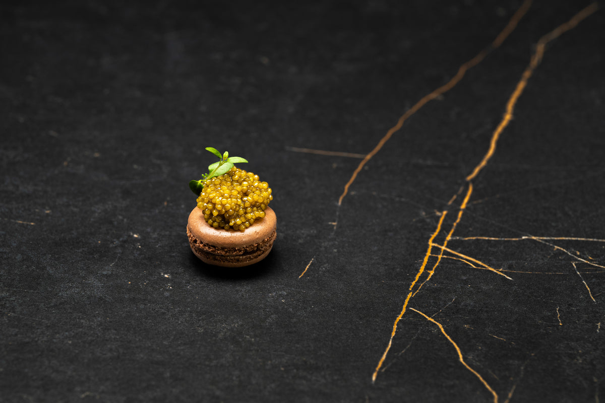 Gourmet chocolate-colored macaron topped with golden caviar pearls and fresh green garnish.
