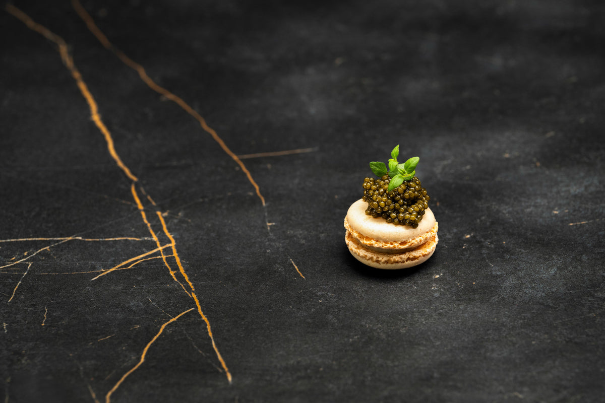 White gourmet macaron topped with premium dark caviar pearls and fresh microgreens on black marble.