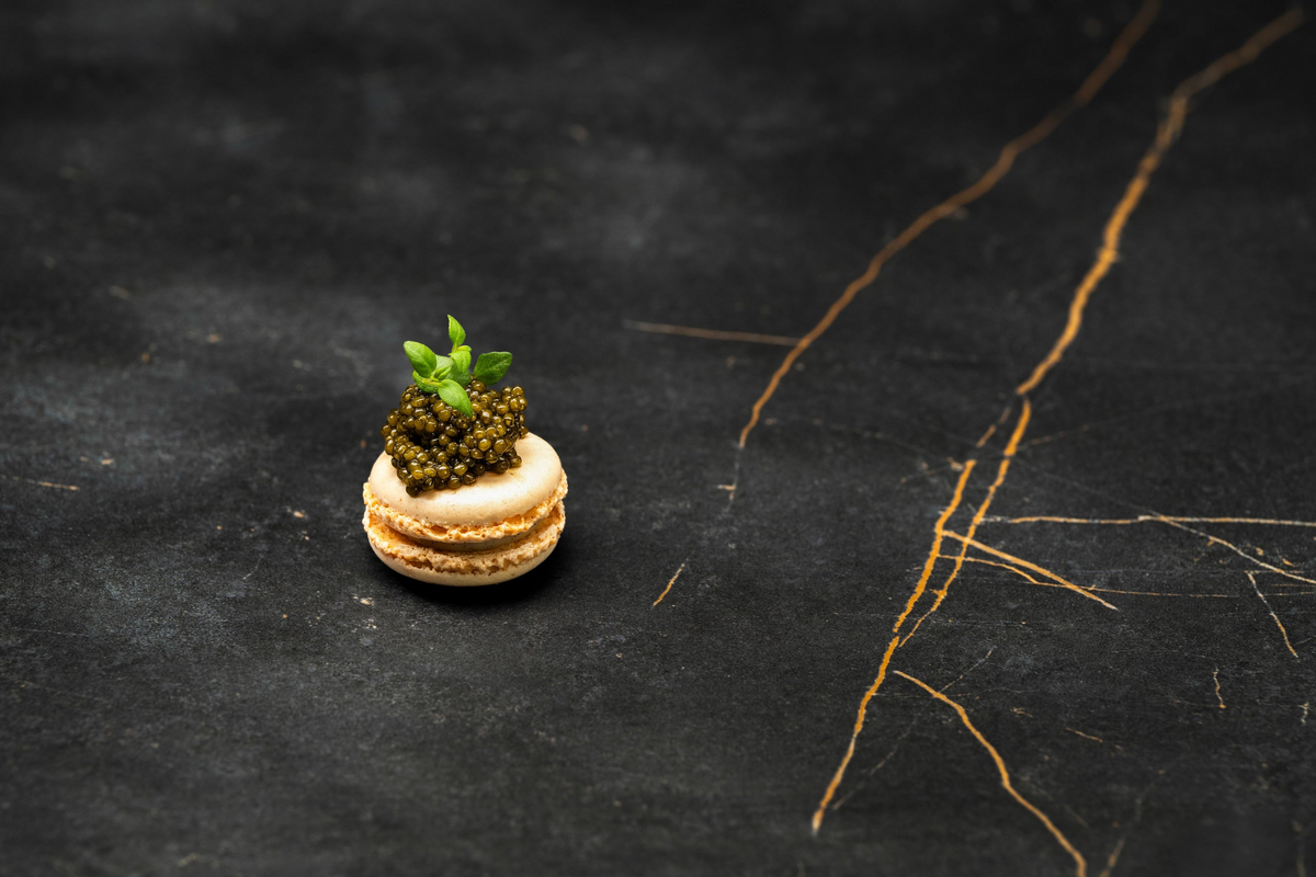 White gourmet macaron topped with premium dark caviar pearls and fresh microgreens on black marble.