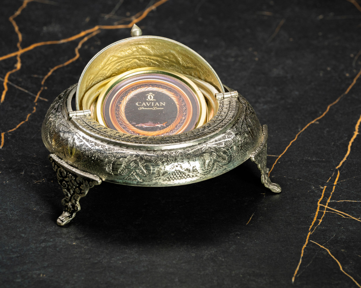 Open silver caviar serving bowl holding a closed Cavian premium caviar tin with black and gold packaging.