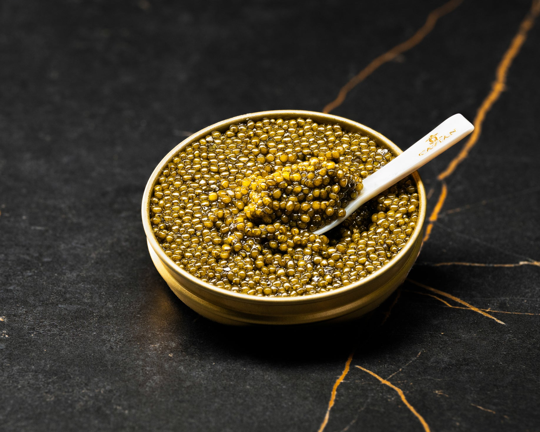 Golden caviar pearls in a gold tin with a mother-of-pearl spoon holding a serving of Cavian Royal Premimu Gold caviar.