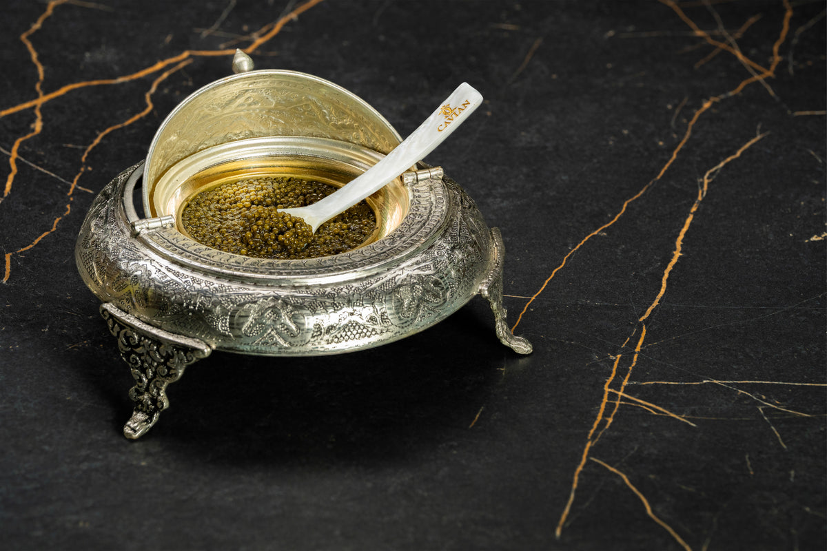 Open engraved silver caviar serving bowl filled with golden caviar and a Cavian mother-of-pearl spoon.