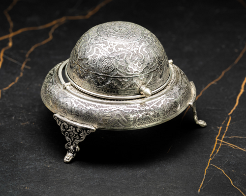 Ornate silver caviar serving bowl with decorative engraved details and a domed lid on black marble.