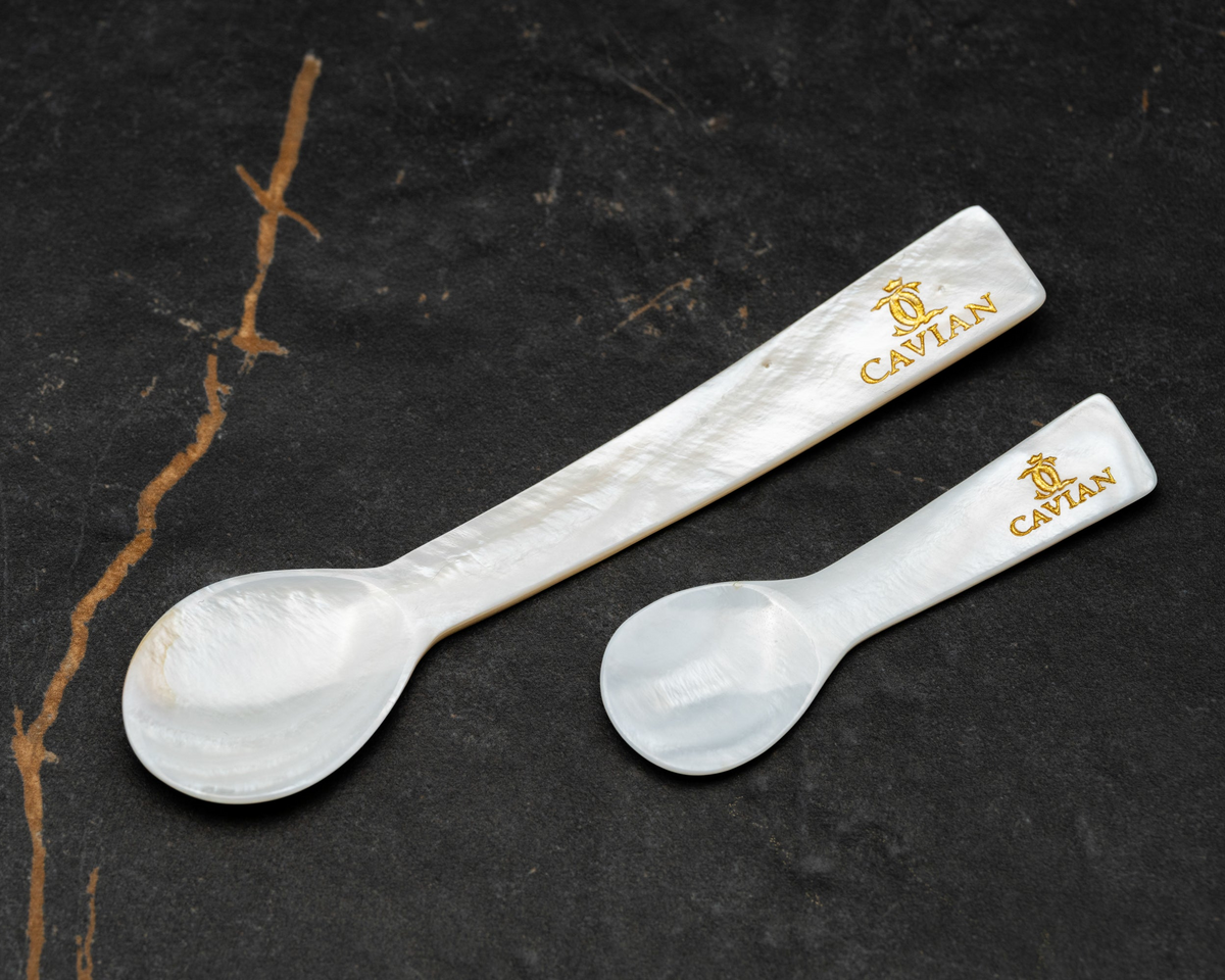 Close-up of two white mother-of-pearl caviar serving spoons in different sizes featuring engraved gold Cavian logos.