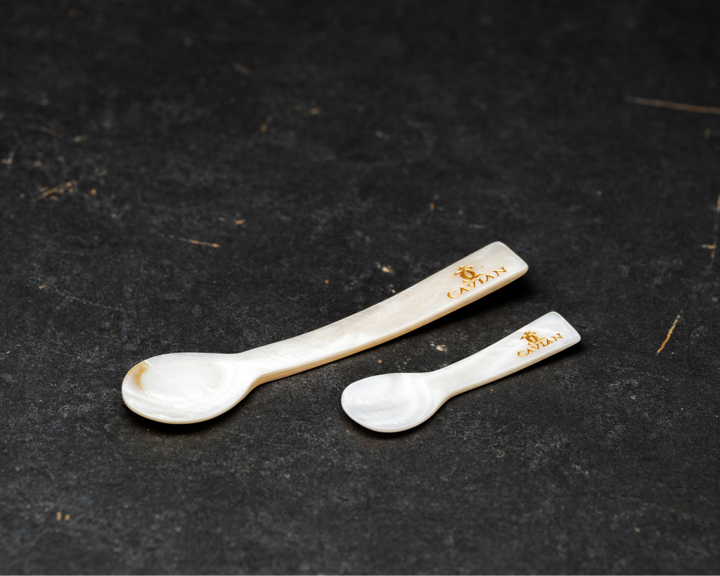 Close-up of two white mother-of-pearl caviar serving spoons in different sizes featuring engraved gold Cavian logos.