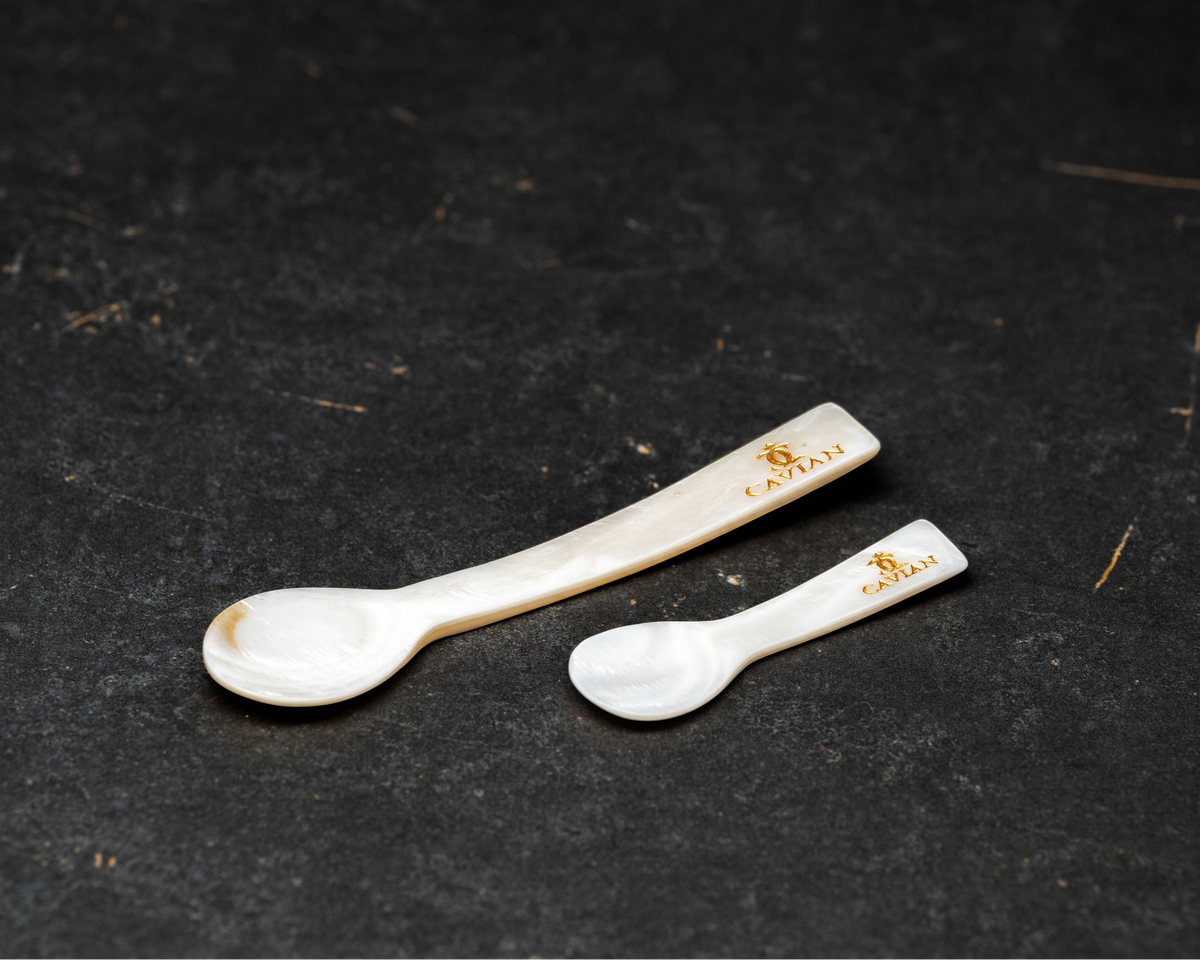 Close-up of two white mother-of-pearl caviar serving spoons in different sizes featuring engraved gold Cavian logos.